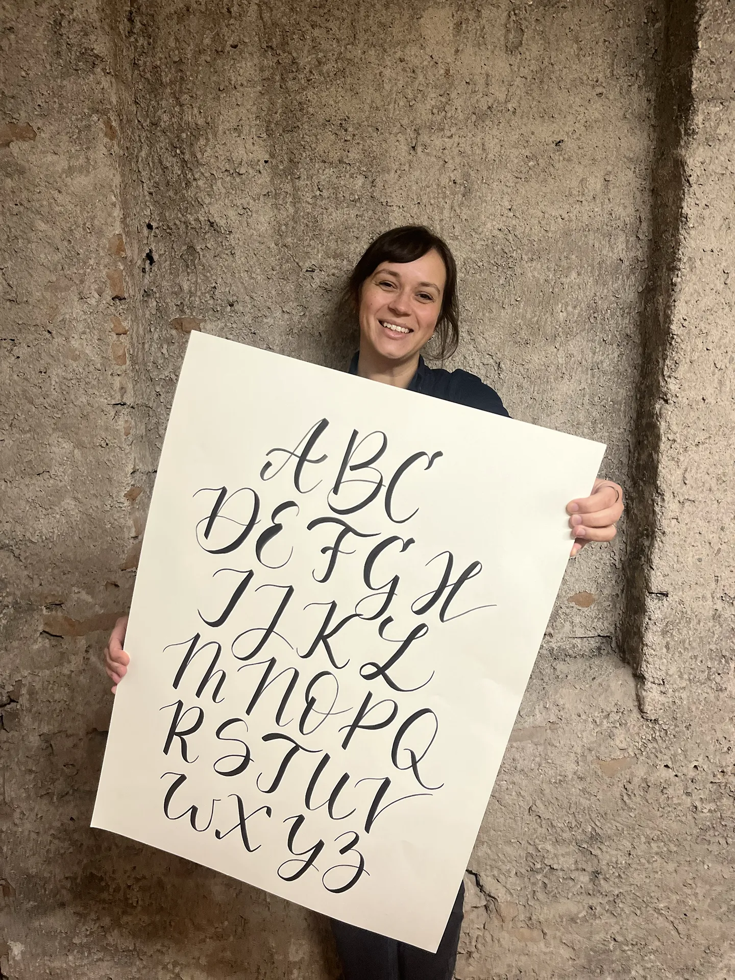 Kamila drží veľký list papiera s ručne písanou abecedou štetcom