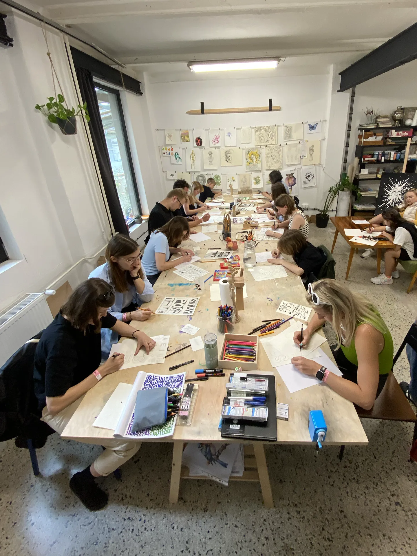 Dlhý stôl v ateliéri, účastníci workshopu cvičia lettering s pomôckami