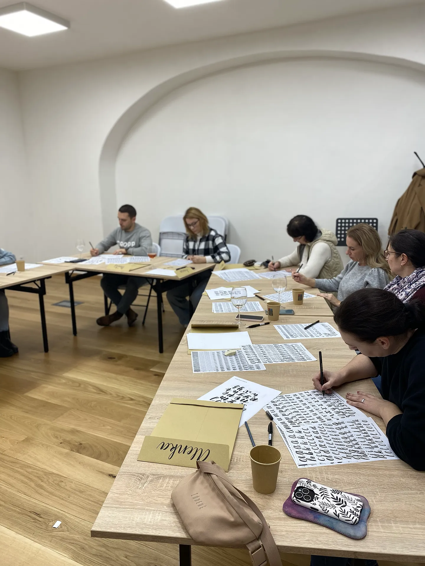 Účastníci sedia pri stoloch v svetlej miestnosti a cvičia lettering na pracovných listoch