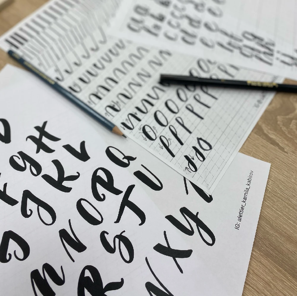 Pracovné listy na brush lettering s čiernou abecedou a cvičnými ťahmi, na stole leží štetec‑fix